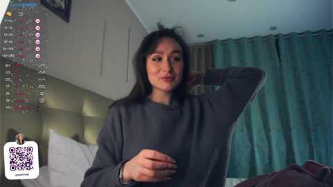 Media: Video of a woman with long black hair, wearing a gray sweater, sitting on a bed with white sheets, behind teal curtains.