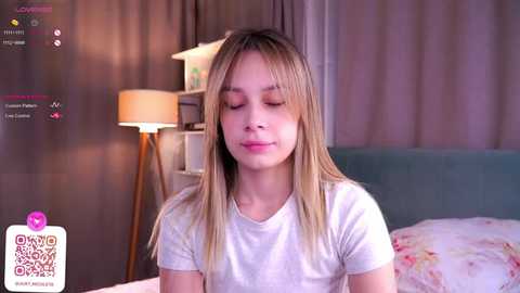 Media: Video of a young, light-skinned girl with straight, blonde hair, wearing a white T-shirt, sitting in a cozy bedroom with a lamp, floral pillow, and beige curtains.