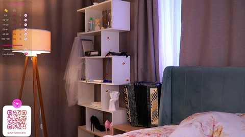 Media: Video of a modern bedroom with a white, cube-shaped shelving unit, a teal headboard, a standing lamp, and a white bed with floral sheets.