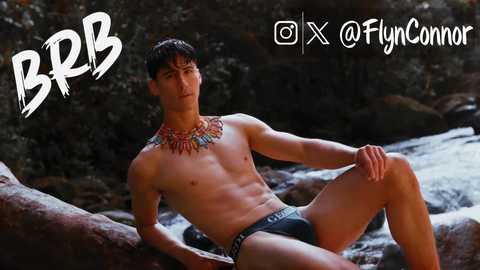 Media: Video of a shirtless, muscular man with short dark hair, wearing only black briefs and a colorful beaded necklace, posing confidently on a large rock beside a stream.