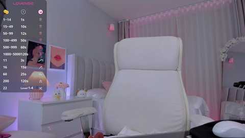 Media: A video of a modern, minimalist spa room with a white massage chair, pink curtains, and a white vanity table with a lamp.