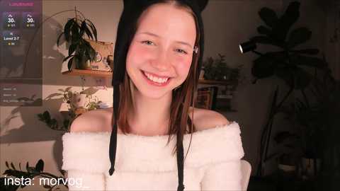 Media: Video of a smiling young woman with fair skin and long brown hair, wearing a white off-shoulder sweater, standing indoors with plants in the background.