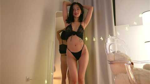 Media: Video of a slim, light-skinned woman with medium-length dark hair, wearing black lace lingerie, standing confidently in a modern, softly lit bedroom.