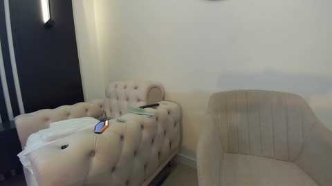 Media: Video of a minimalist hotel room with beige, tufted sofa bed and matching armchair against cream walls, featuring a small side table holding a smartphone and a book, and a modern wall-mounted light fixture.