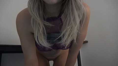 Media: A video of a slender, light-skinned woman with long, straight blonde hair, wearing a purple lace bra and no panties, leaning forward slightly. The background is a plain, light-colored wall.