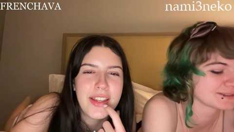 Media: Video of two young women, one with long black hair and light skin, and the other with green hair and fair skin, in a cozy bedroom setting.