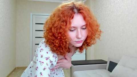 Media: Video of a fair-skinned woman with curly, bright red hair, wearing a floral-patterned robe, gently touching her chest, in a beige-tiled hallway with white doors and a light-colored couch in the background.