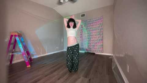 Media: Video of a woman with curly black hair, wearing a white crop top and black patterned pants, dancing joyfully in a sparsely decorated room with gray walls and a colorful abstract mural.