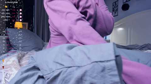 Media: Video of a person in a light pink hoodie, sitting on a bed with messy gray sheets, surrounded by a dark, cluttered room with a TV displaying chat messages.