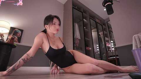 Media: Video of an East Asian woman with medium build, light skin, and dark hair in a black one-piece swimsuit, posing seductively on a wooden floor in a modern, dimly lit room with mirrors and a black lamp.