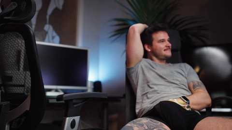 Media: Video of a tattooed, muscular white man in a grey t-shirt and shorts, lounging on a gaming chair, arms raised, in a dimly lit room with a computer monitor and a large plant.