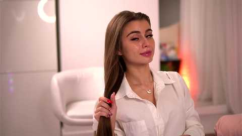 Media: Video of a young, fair-skinned woman with long, straight brown hair, wearing a white button-down shirt, sitting in a modern, white-lit room with a white chair and pink couch in the background.