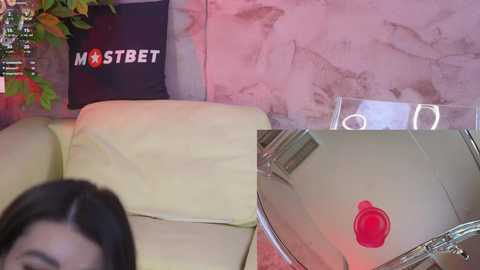 Media: A video featuring a woman with long dark hair, partially visible, sitting on a beige leather couch. A black pillow with \"MOSTBET\" text and a red star is behind her. A metallic red button is attached to her shirt.