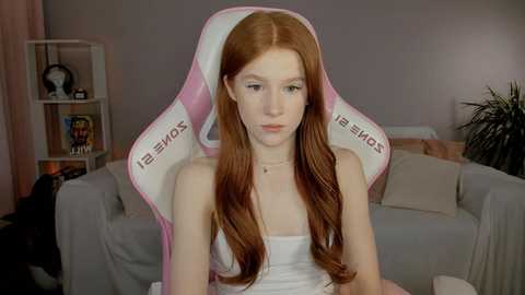 Video of a young, pale-skinned, red-haired woman in a white tank top, sitting in a pink and white gaming chair in a cozy, dimly lit living room with a gray sofa and shelves.