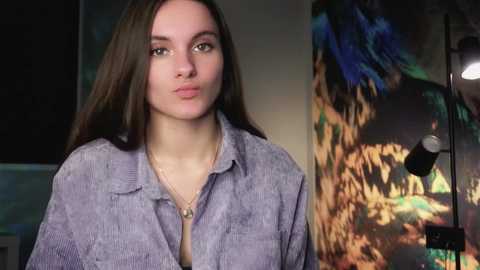 Media: Video of a young woman with long, straight brown hair, wearing a light purple, ribbed, long-sleeved shirt. She has a neutral expression. Background features a colorful, abstract painting with bright hues of blue, orange, and green.