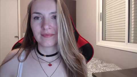 Media: A video of a young woman with long blonde hair, fair skin, and a neutral expression, wearing a black choker, multiple necklaces, and a red pendant, seated in a gaming chair in a modern room with white blinds and gray-patterned carpet.