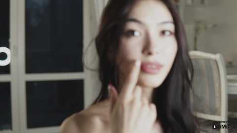 Media: A close-up video of a young woman with long, dark hair, wearing a pink dress, standing in a brightly lit room with a window. She's covering her mouth with her fingers.