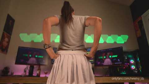 Media: Video of a muscular woman with dark hair in a ponytail, wearing a gray shirt and white skirt, standing with hands on hips in a modern gaming room with multiple monitors and colorful LED lights.