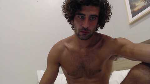 Media: Video of a shirtless, muscular Middle Eastern man with curly brown hair, wearing a contemplative expression, sitting on a bed with white sheets in a minimalist bedroom with a framed landscape painting on the wall.