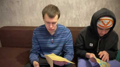 Media: Video of two Caucasian boys, one in a striped blue shirt, the other in a black hoodie, both focused on a yellow paper airplane on a brown couch against a textured gray wall.