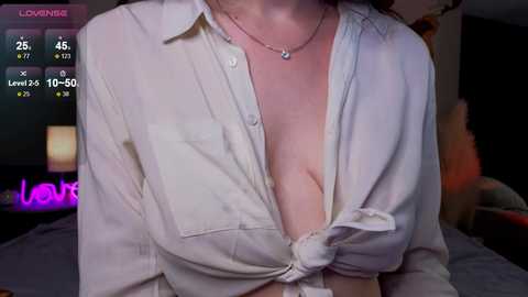 A video of a woman with fair skin, wearing an unbuttoned white shirt revealing her cleavage. She has a delicate silver necklace. The background is dimly lit, with indistinct furniture and a blurred figure.