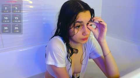 Media: Video of a young woman with wet, dark hair, wearing a white, wet T-shirt, in a white bathtub, looking sad.