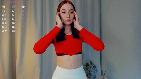 Media: Video of a fair-skinned, red-haired woman with long straight hair, wearing a red crop top and white high-waisted pants, adjusting her hair. Background features a gray curtain and a small potted plant.