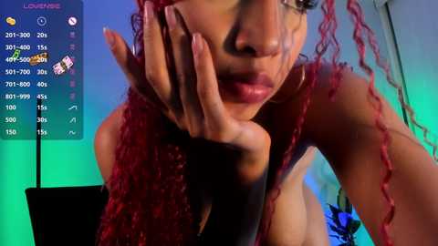 Media: Video of a young woman with medium brown skin and vibrant red curly hair. She is topless, with her hand covering her face, wearing a black glove. Background shows a green and blue-lit room with a video game interface on the left.