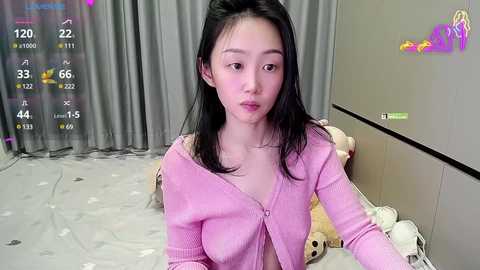 Media: A video of an East Asian woman with straight black hair and light skin, wearing a pink cardigan, sitting on a bed with plush toys, in a room with gray curtains and a TV showing a live stream.