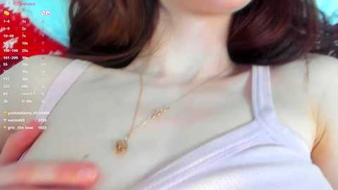 Media: Video of a fair-skinned woman with long brown hair, wearing a white tank top, and a delicate gold necklace. Background includes a red and blue patterned fabric.