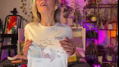 Media: Video of an older woman with blonde hair, wearing a white Canada T-shirt, gesturing animatedly while holding a map. Background features colorful plants, a chair, and a colorful painting, with warm ambient lighting.