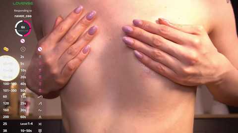 Media: A close-up video of a fair-skinned, topless person with light purple nail polish, covering their small breasts with both hands. The background is blurred, showing a digital screen with a 'Live' status and a circular gauge.