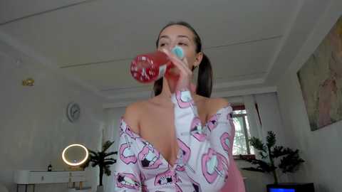 Media: Video of a young woman with light skin and dark hair in pigtails, wearing a pink off-shoulder blouse, drinking from a red and white striped straw, in a modern, bright room with white walls and potted plants.