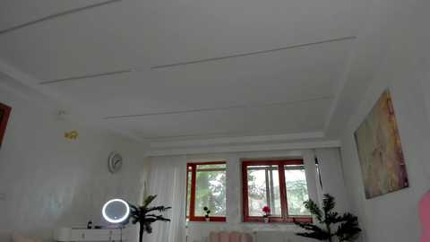 Media: Video of a minimalist, bright living room with white walls, a large window, and a round clock. A selfie ring light is placed on the floor, and a colorful abstract painting hangs on the wall.