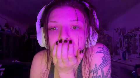 Media: Video of a young woman with wet hair, wearing headphones, covering her mouth with her hand. She has a tattoo on her left shoulder. The background is dimly lit with purple lighting, giving an eerie ambiance.