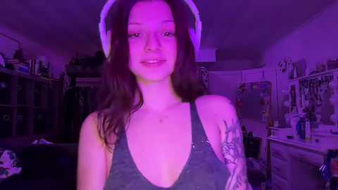 Media: A video of a young woman with long, dark hair wearing a black tank top, standing in a dimly lit room with purple lighting. She has tattoos on her left arm.