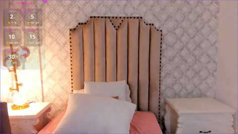 Media: Video of a stylish, modern bedroom with a beige upholstered headboard, white pillows, a white nightstand, and patterned wallpaper.