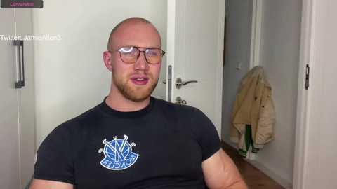 Media: Video of a muscular, bald, fair-skinned man with a beard, wearing glasses and a black \"Firefighters\" t-shirt, standing indoors near a door with a beige jacket hanging.