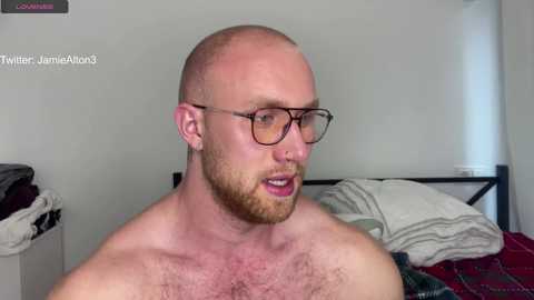 Media: A video of a bald, muscular, fair-skinned man with a beard and glasses, standing shirtless in a bedroom with white walls, a bed with messy sheets, and a partially visible white window.