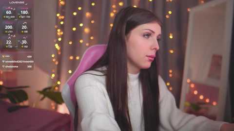 Media: A video of a young woman with long, straight brown hair, wearing a white sweater, seated in a pink chair, looking contemplative in a softly lit, cozy room with fairy lights and a bed in the background.