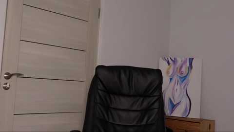 Media: Video of a minimalist office corner with a closed beige wooden door, a black leather chair, and a vibrant abstract painting on a light gray wall.