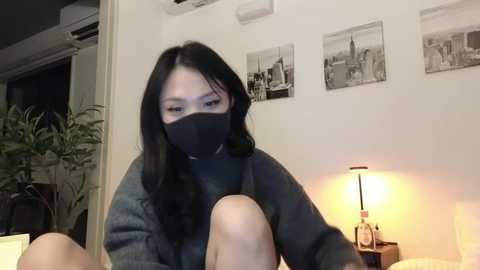 Media: Video of an East Asian woman with long black hair, wearing a black face mask, grey sweater, and sitting on a bed. Background features a white wall with framed black-and-white photos and a lamp.