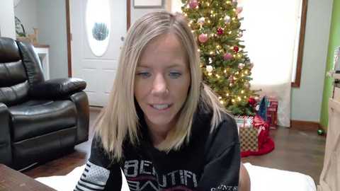 Media: Video of a blonde woman in a black sweater, smiling, in a cozy living room with a decorated Christmas tree and wrapped gifts.