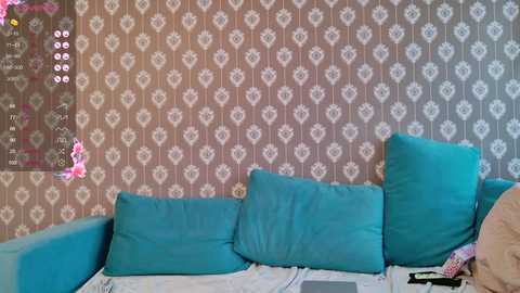 Media: Video of a vintage-style living room with a teal sofa against a patterned wallpaper, featuring a pink and white floral design. The room is cluttered with a phone and a pink object on the right.