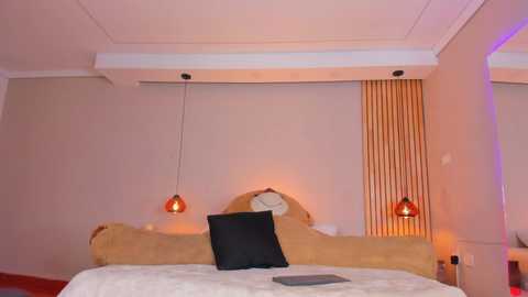 Media: Video of a modern, minimalist bedroom with beige walls, a wooden headboard, and two red pendant lights. A black pillow and a laptop rest on a white bedspread.