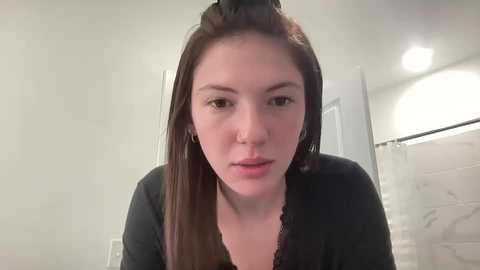 Media: A young woman with straight, long brown hair is seen from the chest up in a bathroom with white walls and a shower curtain. She has a neutral expression, wearing a black top.