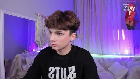 Media: Video of a young Caucasian boy with short brown hair, wearing a black \"ATI\" t-shirt, sitting on a light-colored couch in a dimly lit room with purple and white lighting.