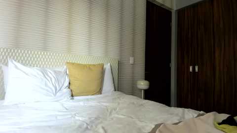 Media: Video of a modern bedroom with a white bed featuring a textured headboard, white linens, and a mustard-yellow pillow. The background includes vertical, striped wallpaper and a dark wooden wardrobe door.