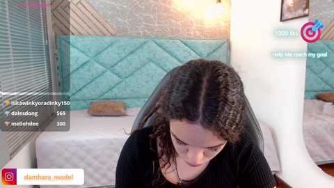 Media: Video of a young woman with curly hair, wearing a black top, seated in a cozy, teal-upholstered room, with text overlays indicating her social media presence.