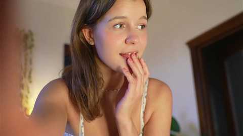 Media: Video of a young Caucasian woman with fair skin, straight brown hair, and green eyes, wearing a white tank top, thoughtfully biting her fingernails. Background shows a dimly lit room with a wooden door and a string of fairy lights.